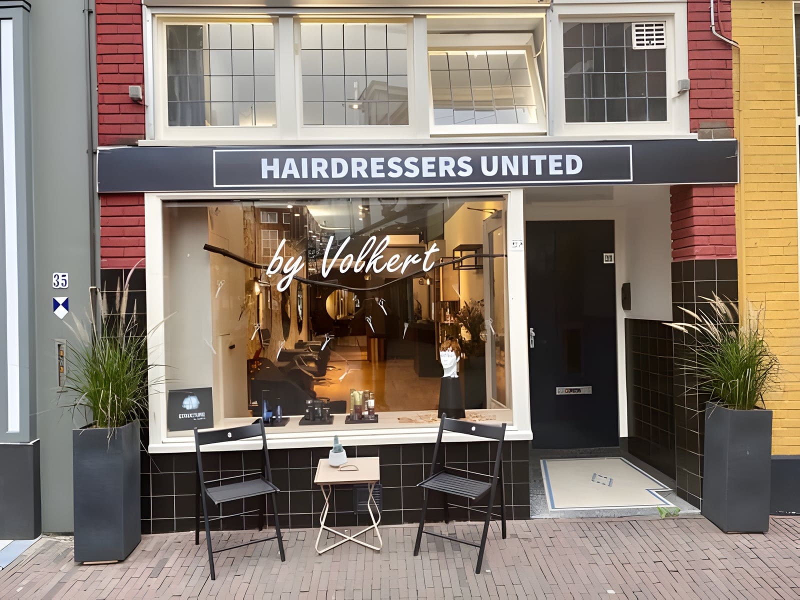 De gevel van Hairdressers United by Volkert met het zwarte uithangbord en twee stoeltjes bij de etalage