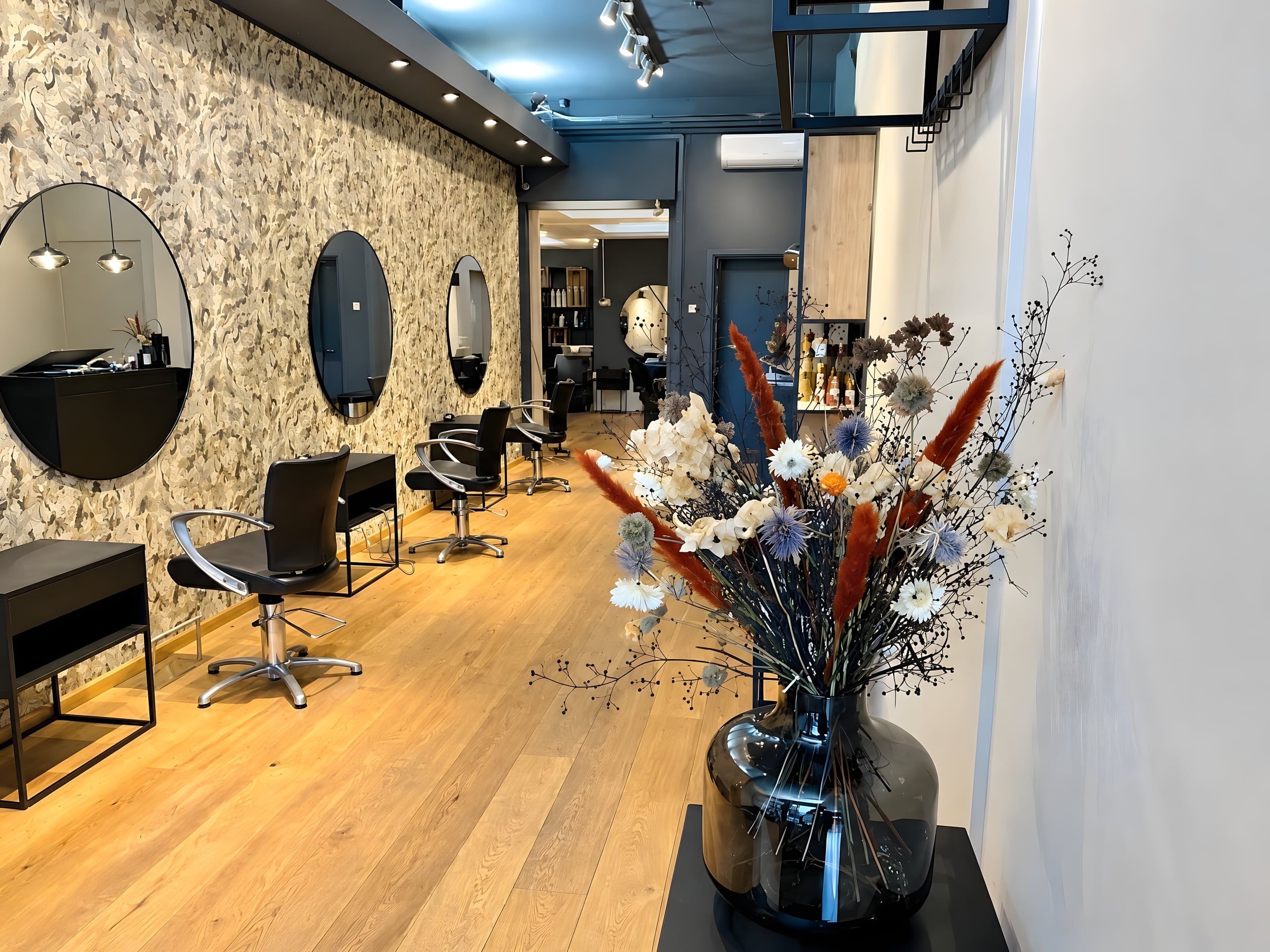 De salon van Hairdressers United: zwarte stoelen en ronde spiegels langs een wand met botanisch motief, een eikenhouten vloer en een grote vaas met droogbloemen